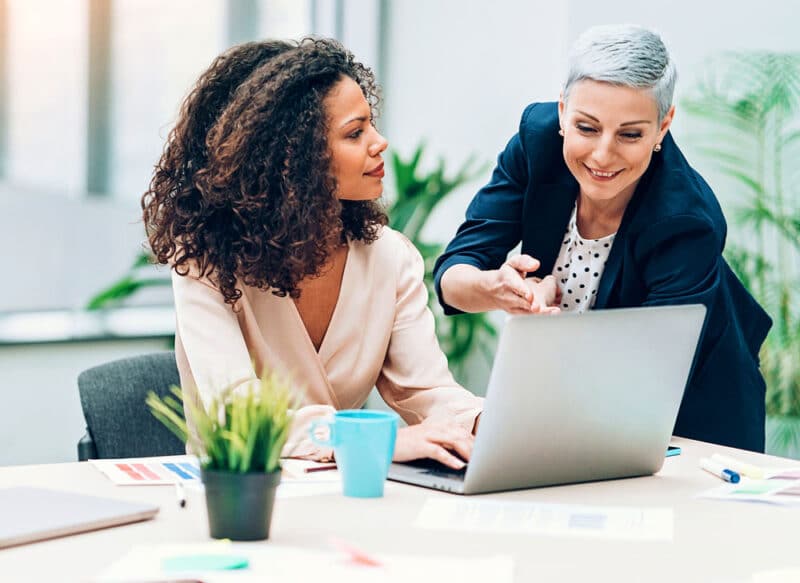 smiling woman explaining to sitting woman while looking at laptop