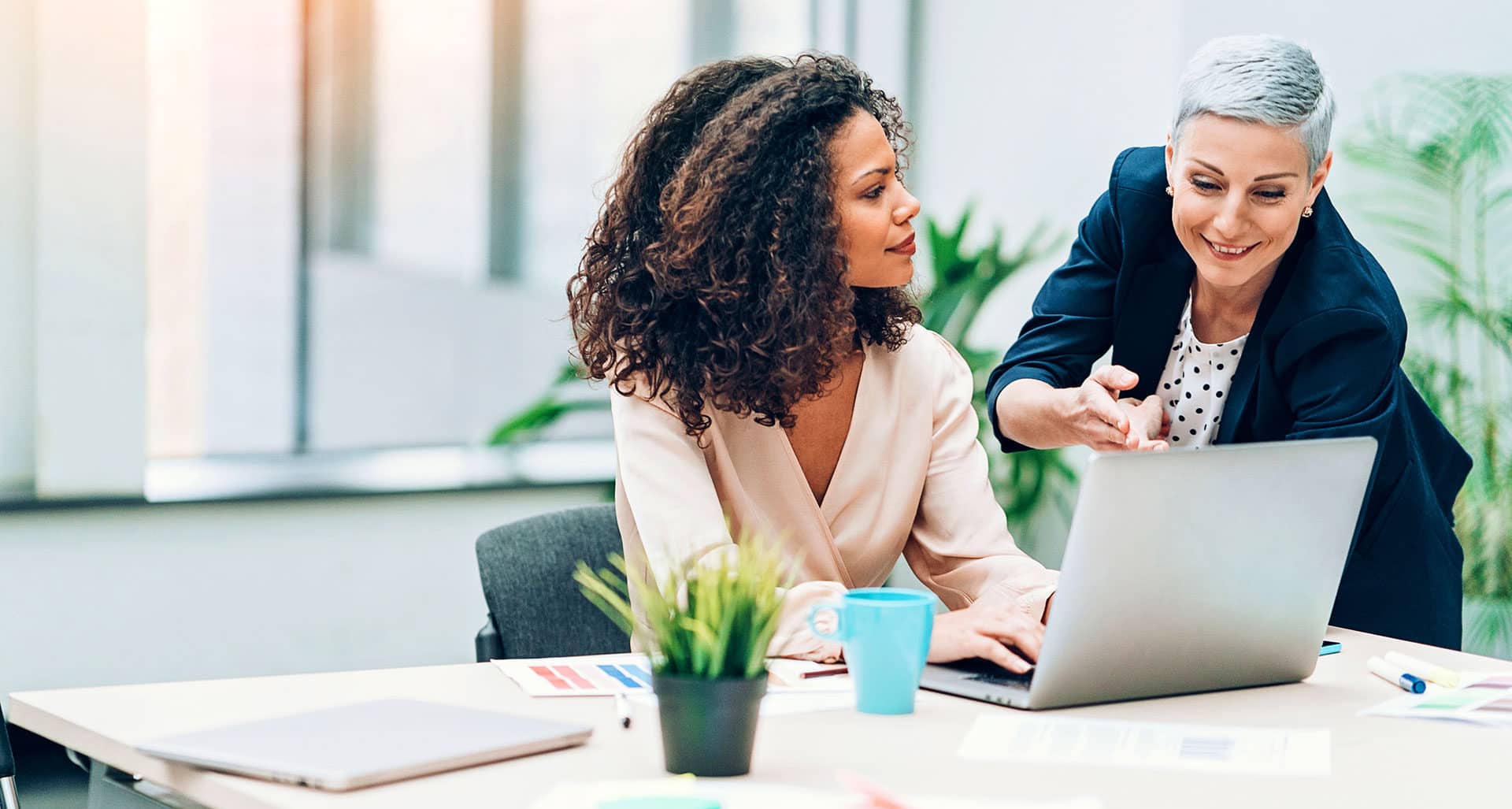smiling woman explaining to sitting woman while looking at laptop